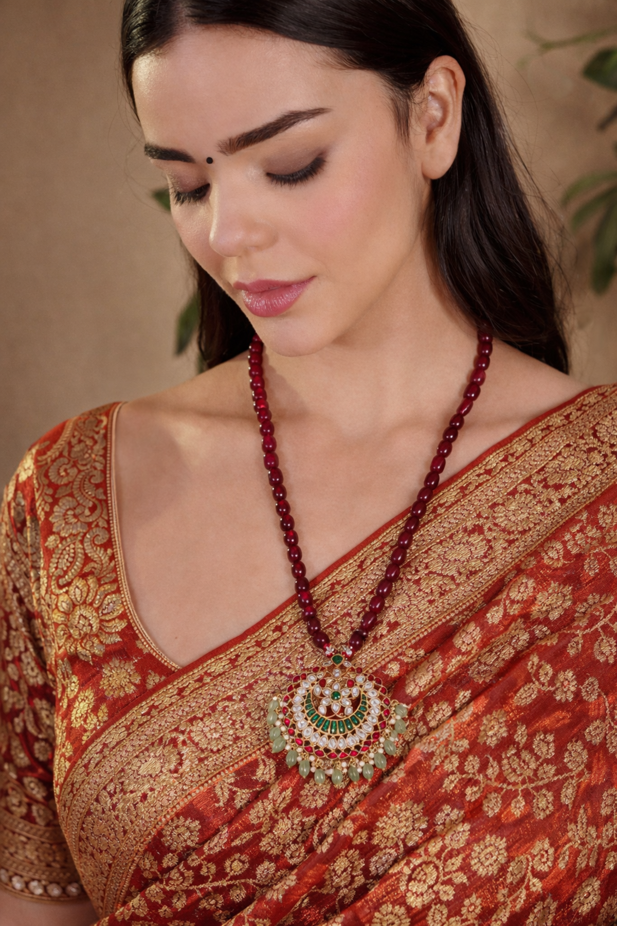 A woman wearing a traditional Indian red and gold saree, featuring a prominent red beaded necklace with a large circular green and gold pendant.