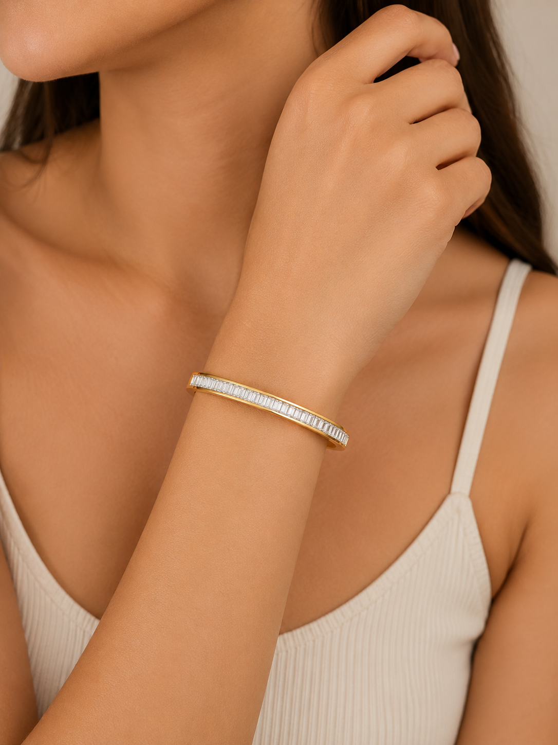 A close-up of a woman wearing a slim gold bangle bracelet with a row of clear pavé-set stones, paired with a white ribbed camisole top.