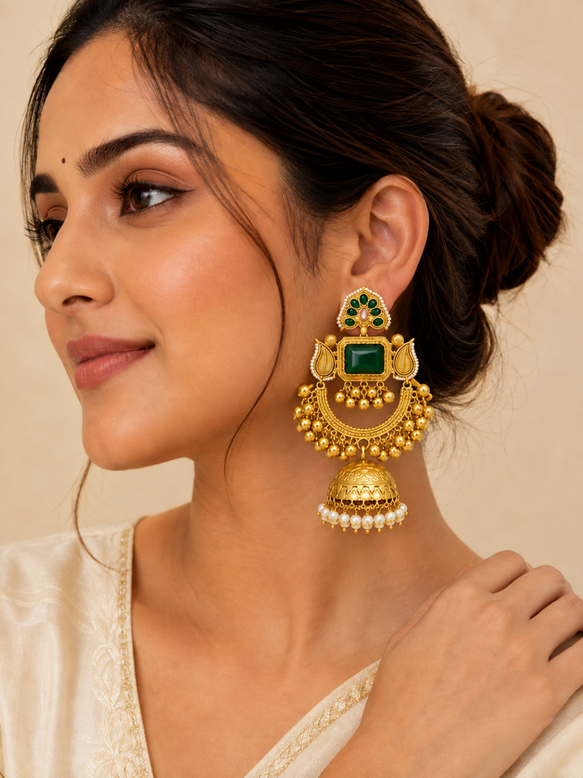 A woman wearing a large traditional Indian gold jhumka earring with a green rectangular stone, gold filigree, and a pearl tassel, shown in a close-up profile shot against a warm beige background.