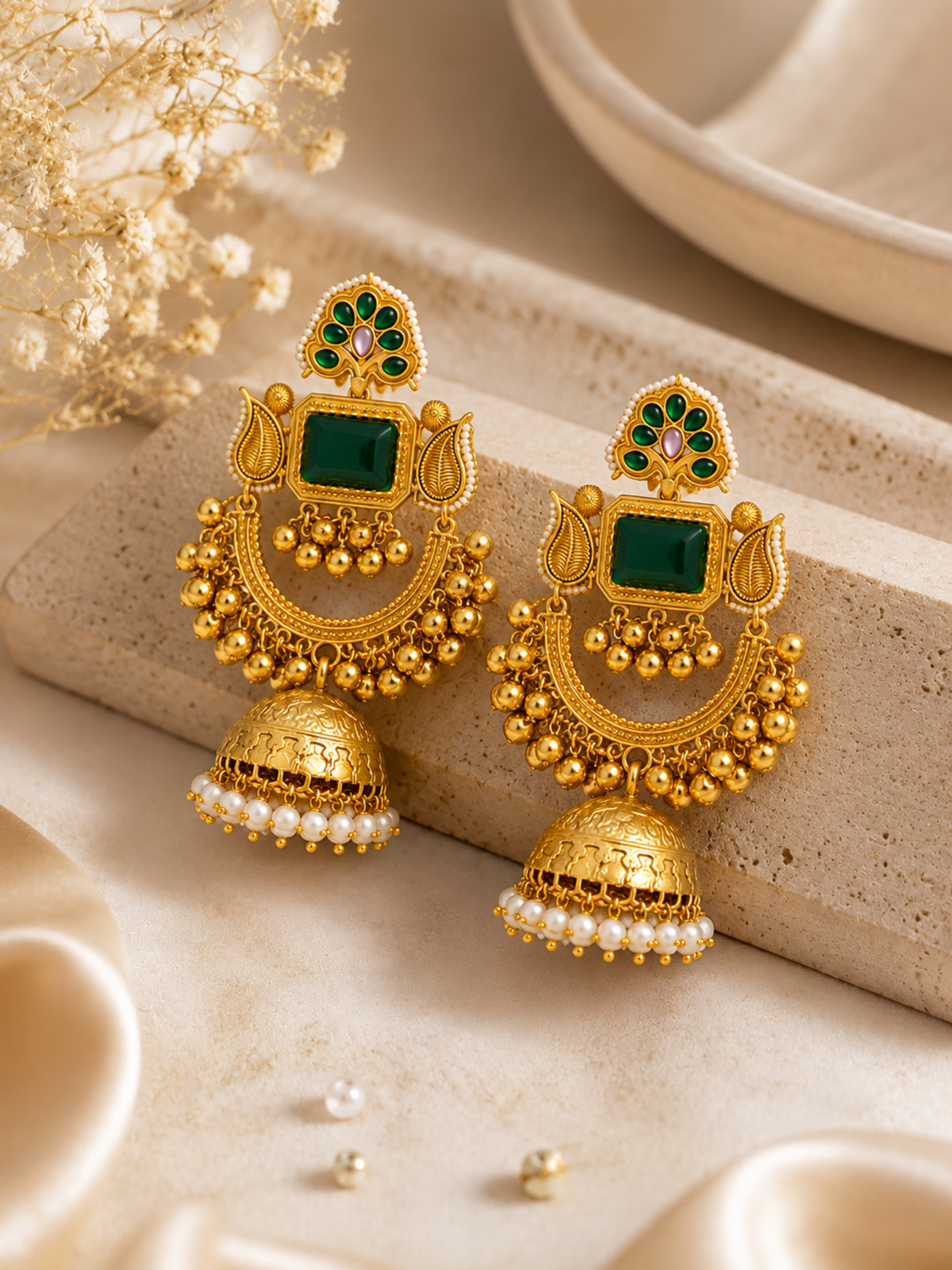 A pair of ornate gold-toned traditional Indian jhumka earrings with green rectangular stones, small green floral accents, and rows of white pearl-like beads at the bottom, displayed on a textured beige stone block with dried flowers in the background.
